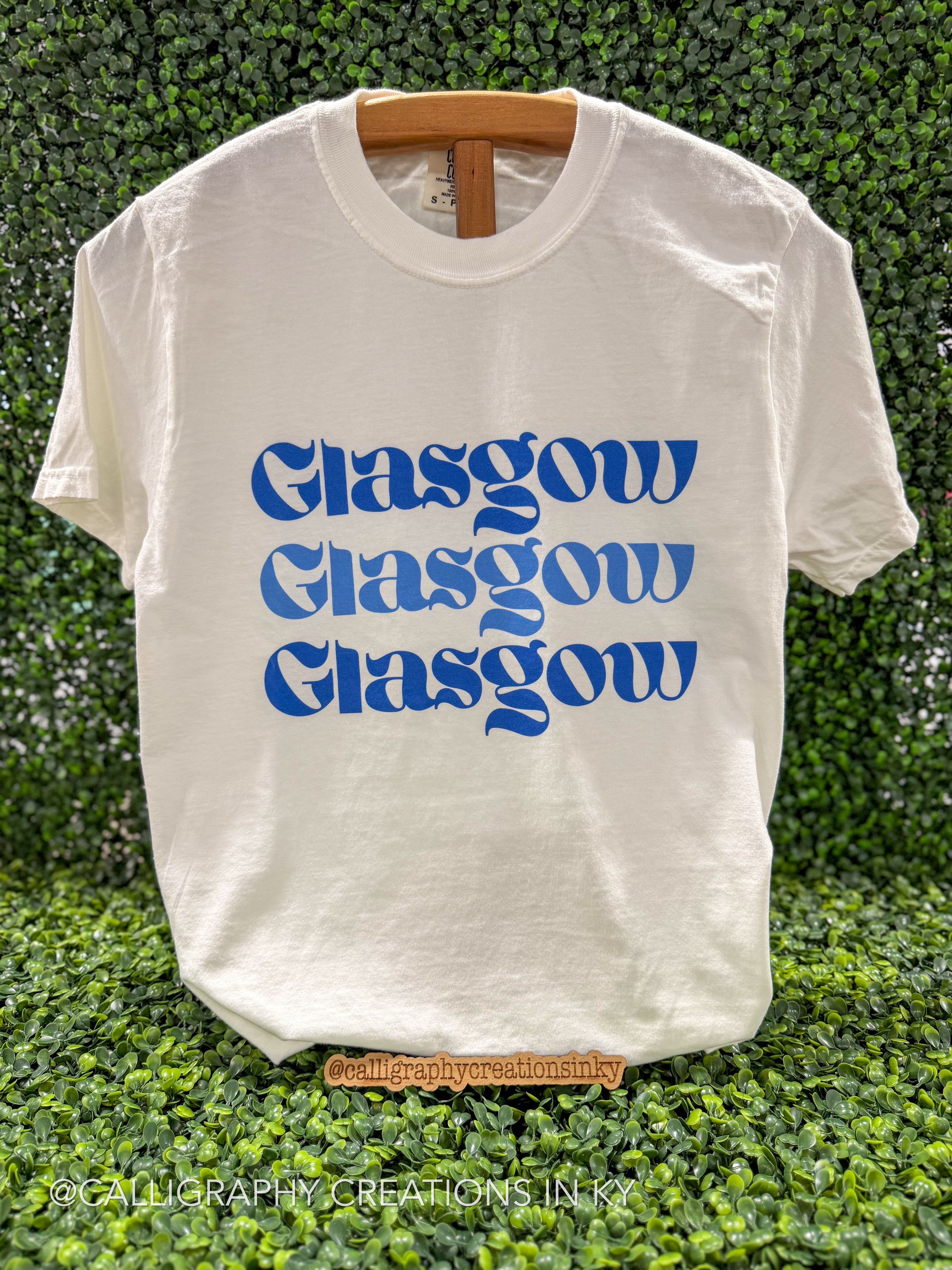 An off-white short-sleeve t-shirt with 'Glasgow' printed three times in blue retro-style lettering, displayed on a wooden hanger against a green hedge background.