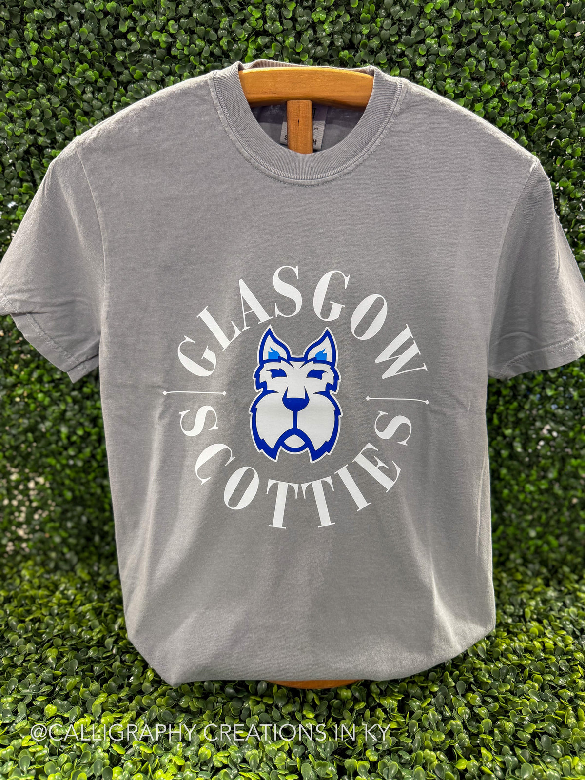A light gray crewneck t-shirt hanging on a wooden hanger against a green hedge background. The shirt features a graphic print of a blue and white Scottish terrier dog inside a circle with the words 'GLASGOW SCOTTIES' in white.