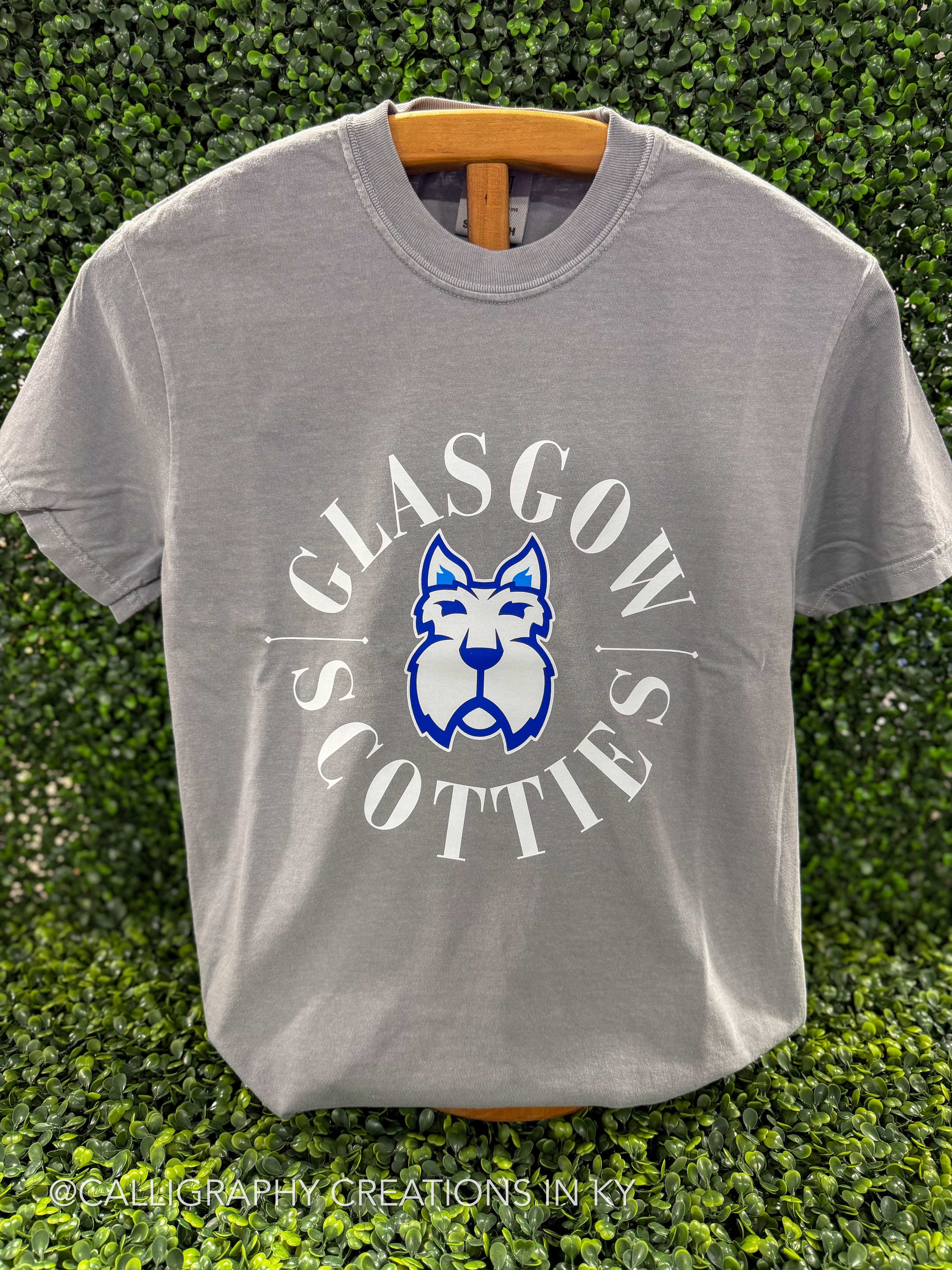 A light gray crewneck t-shirt hanging on a wooden hanger against a green hedge background. The shirt features a graphic print of a blue and white Scottish terrier dog inside a circle with the words 'GLASGOW SCOTTIES' in white.