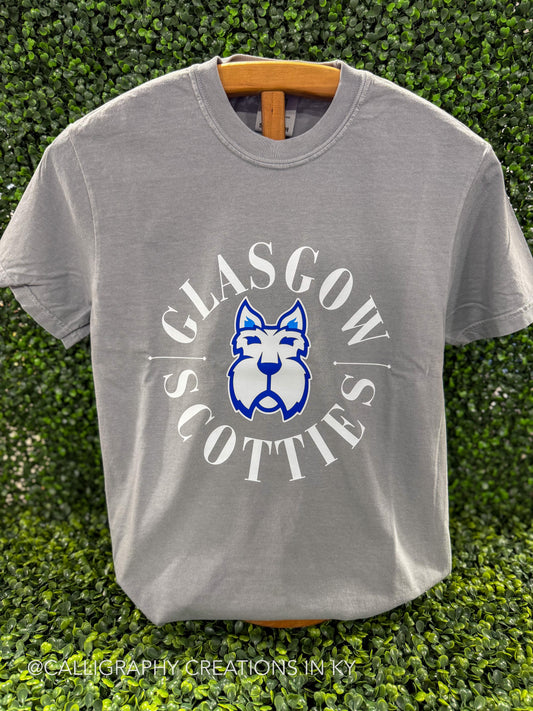 A light gray crewneck t-shirt hanging on a wooden hanger against a green hedge background. The shirt features a graphic print of a blue and white Scottish terrier dog inside a circle with the words 'GLASGOW SCOTTIES' in white.