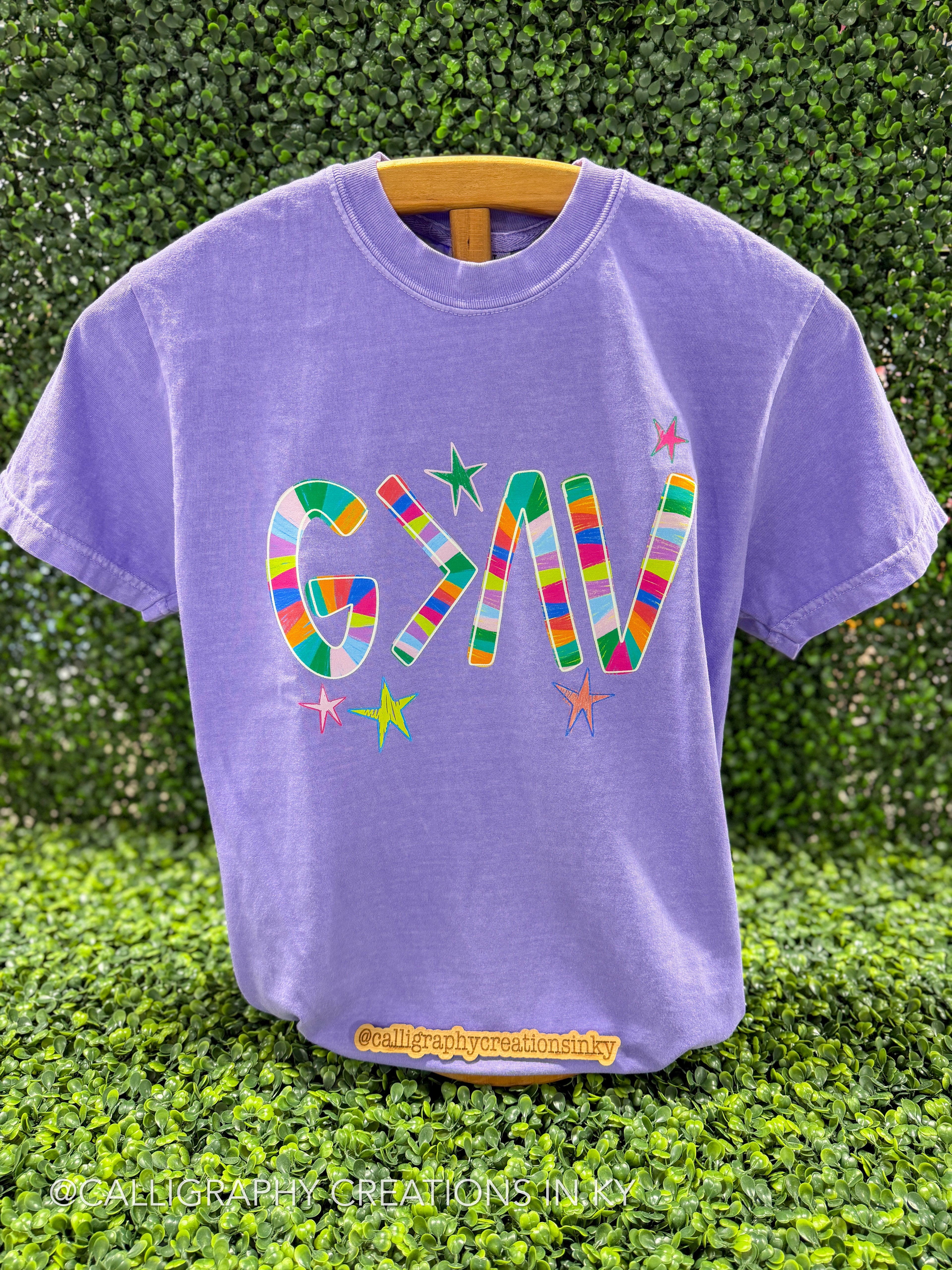 A purple t-shirt with a colorful striped "God Is Greater Than The Highs And Lows" symbol graphic and small stars on the front, displayed on a wooden hanger against a green hedge background.