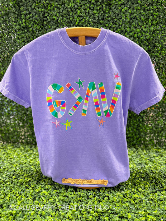 A purple t-shirt with a colorful striped "God Is Greater Than The Highs And Lows" symbol graphic and small stars on the front, displayed on a wooden hanger against a green hedge background.
