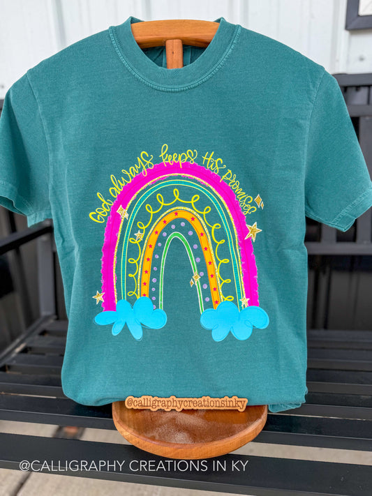 A teal short-sleeve t-shirt displayed on a wooden hanger and stand, featuring a colorful rainbow graphic with the text “God always keeps his promises' in yellow script. The background shows a black slatted outdoor bench.