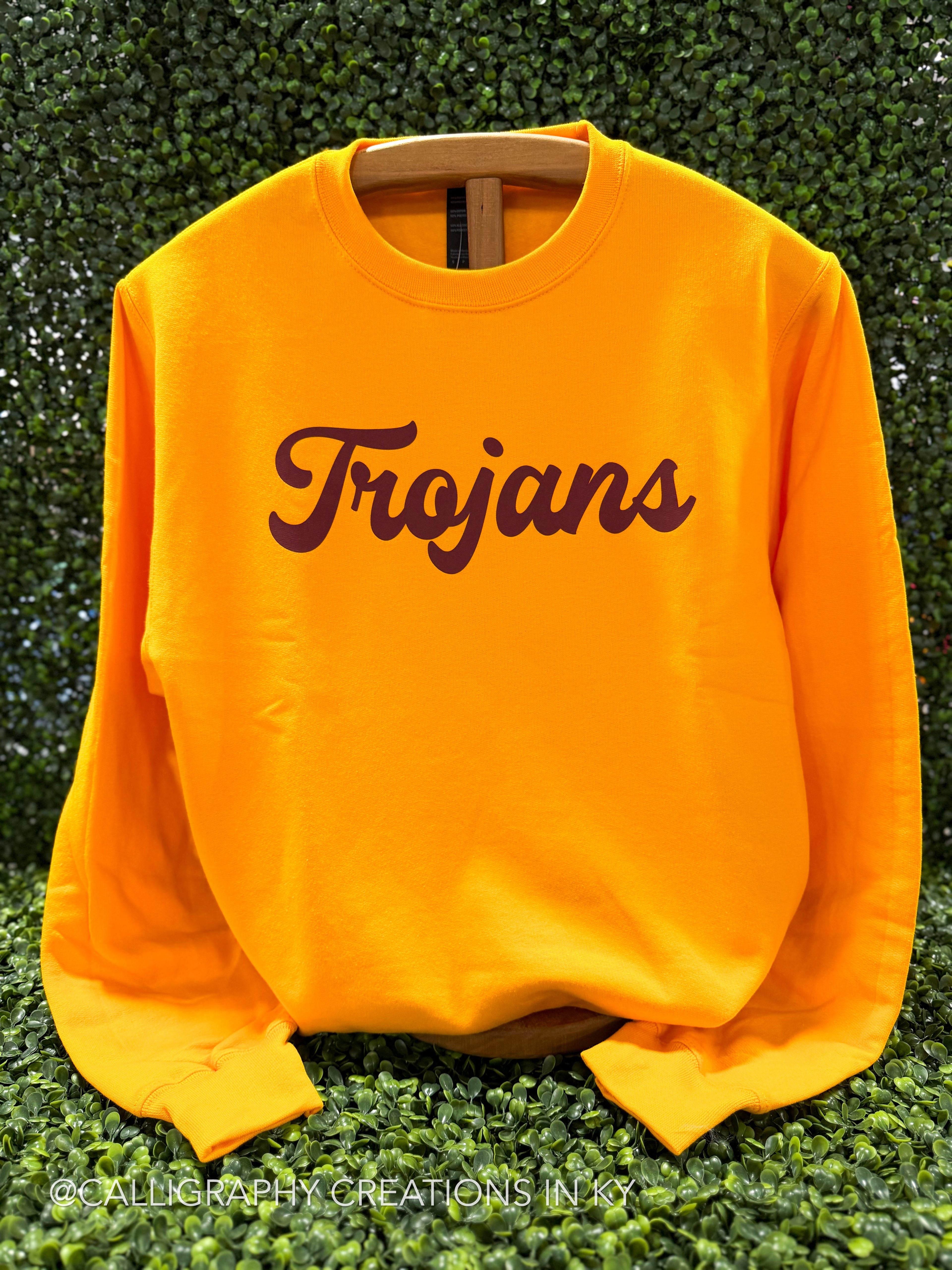 A bright yellow crewneck sweatshirt with 'Trojans' printed across the chest in dark brown script, displayed on a wooden hanger against a green leafy hedge background.