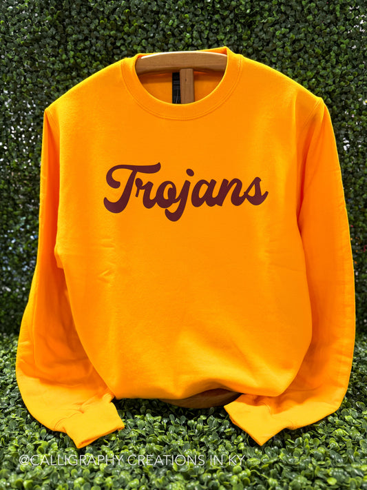 A bright yellow crewneck sweatshirt with 'Trojans' printed across the chest in dark brown script, displayed on a wooden hanger against a green leafy hedge background.