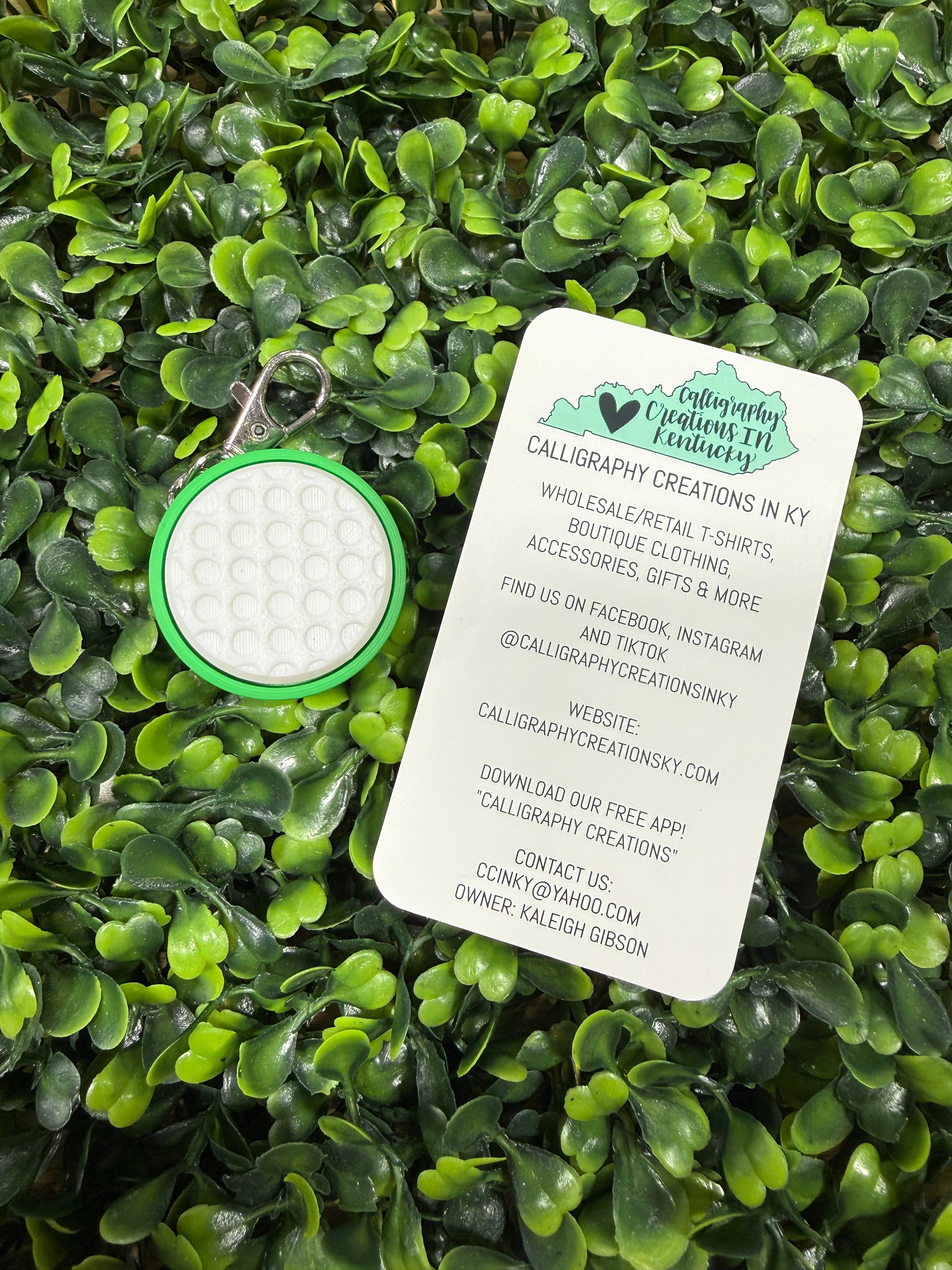 A circular golf-themed keychain with a green border and white textured center, shown next to a white printed business card on a dense green leaf background.