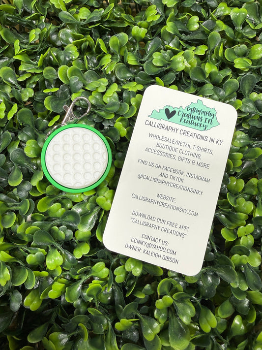 A circular golf-themed keychain with a green border and white textured center, shown next to a white printed business card on a dense green leaf background.