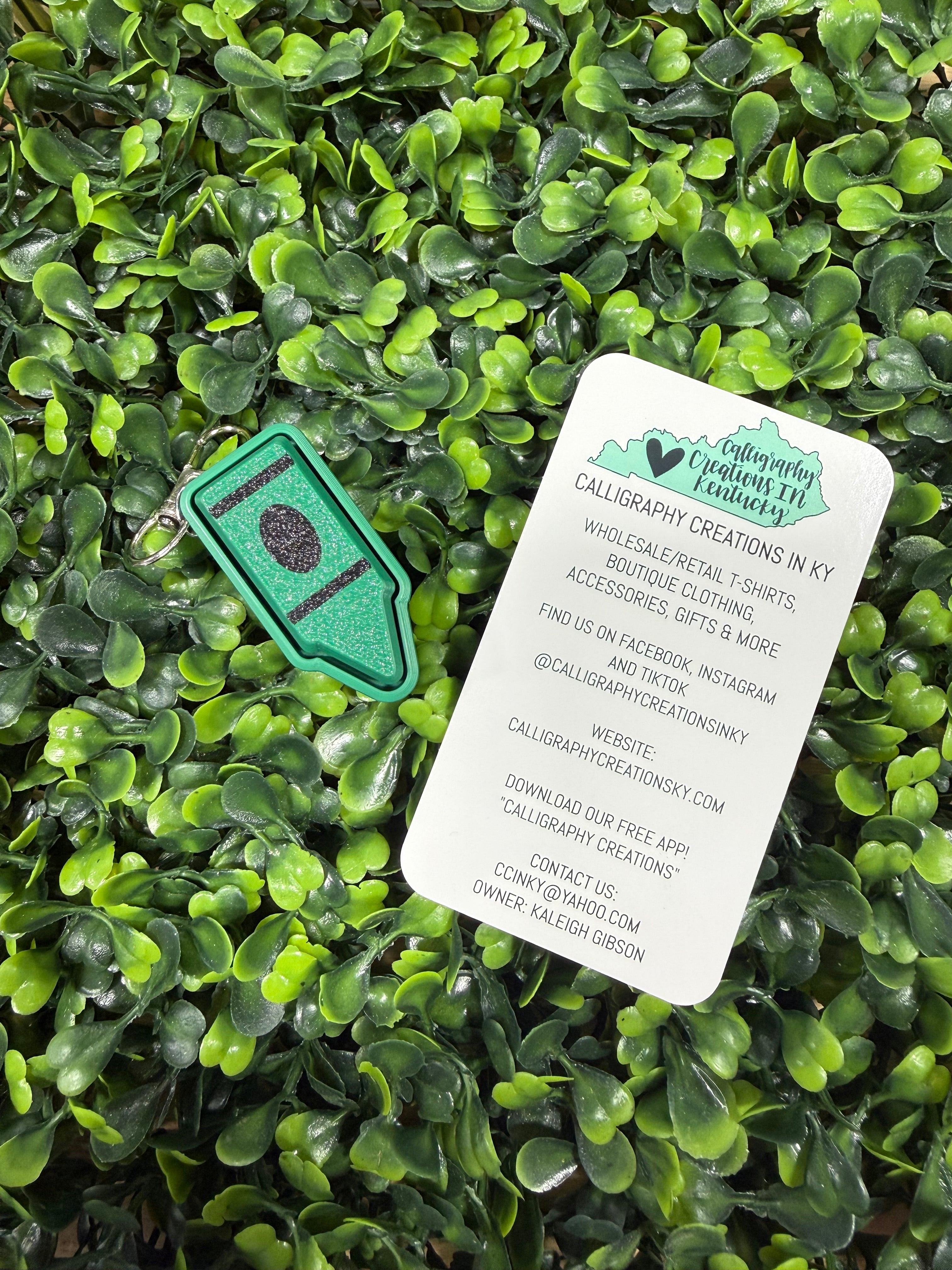 A small green crayon clicker charm with a dark purple circular detail lies on a bed of artificial green leaves, next to a white paper tag with contact information for Calligraphy Creations in KY.