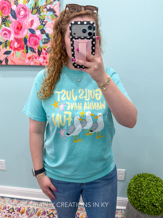 A woman takes a mirror selfie wearing a turquoise graphic t-shirt with seagulls and the text 'Gulls Just Wanna Have Fun' in yellow and white. The background shows a light blue wall with a floral painting and a small green plant.
