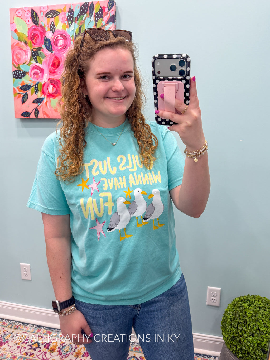 A woman takes a mirror selfie wearing a light blue graphic t-shirt with yellow text and seagull illustrations. The background shows a light blue wall with a floral painting, outlets, and a small potted plant.