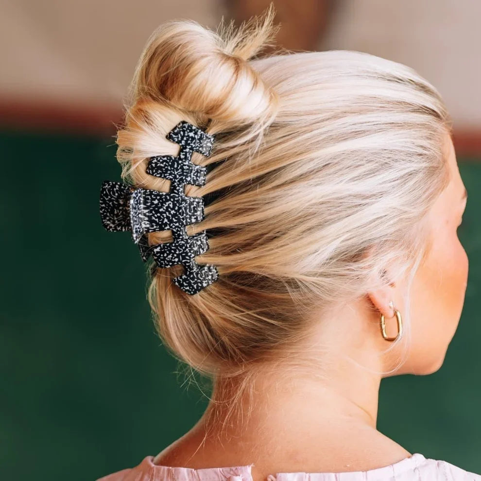 A close-up side view of a woman with blonde hair styled in a messy bun, secured with a large black claw hair clip She is wearing a small gold hoop earring, and the background is a soft, out-of-focus dark green.