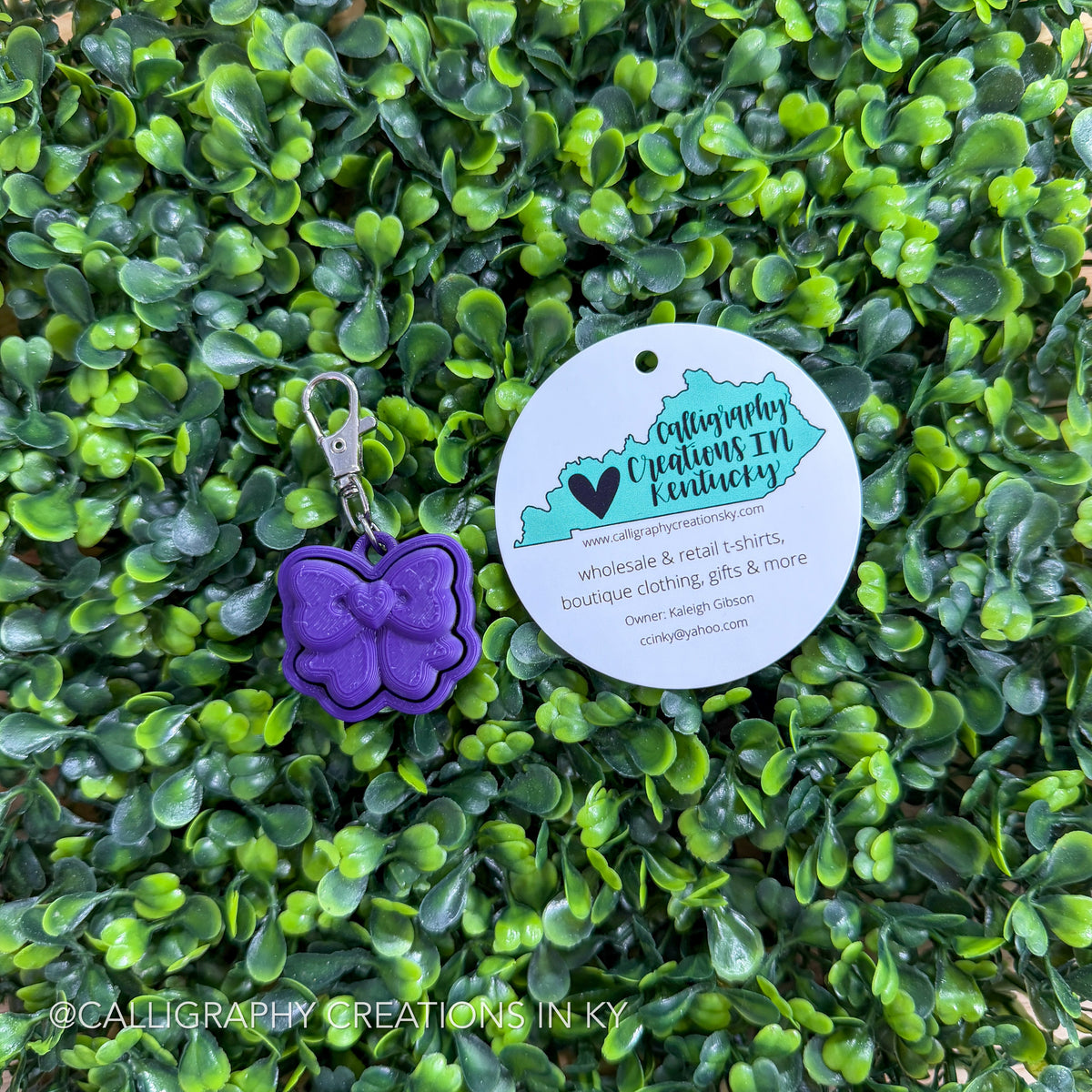 Purple bow shaped fidget clicker keychain with a tag on a green leafy background