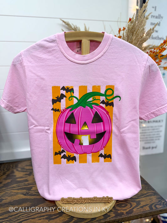 A light pink short-sleeve t-shirt on a wooden hanger, featuring a graphic of a jack-o'-lantern with a pink striped pumpkin body, green stem, and black bat silhouettes on an orange striped background.