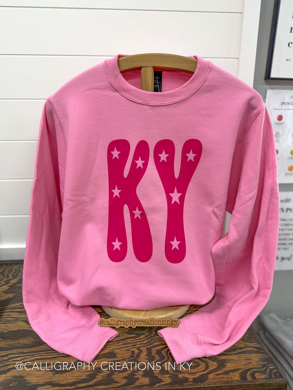 A pink crewneck sweatshirt with a large retro-style 'KY' graphic in dark pink with white stars, displayed on a wooden hanger on a wooden table.
