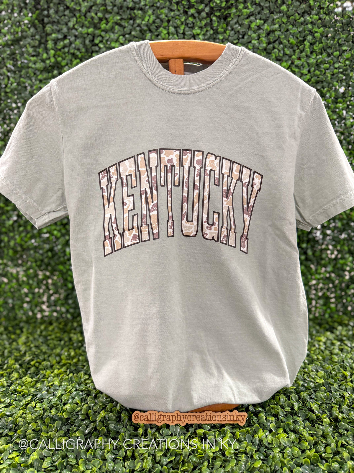 Sage green shirt with Camo ‘Kentucky’ across the chest on a wooden stand