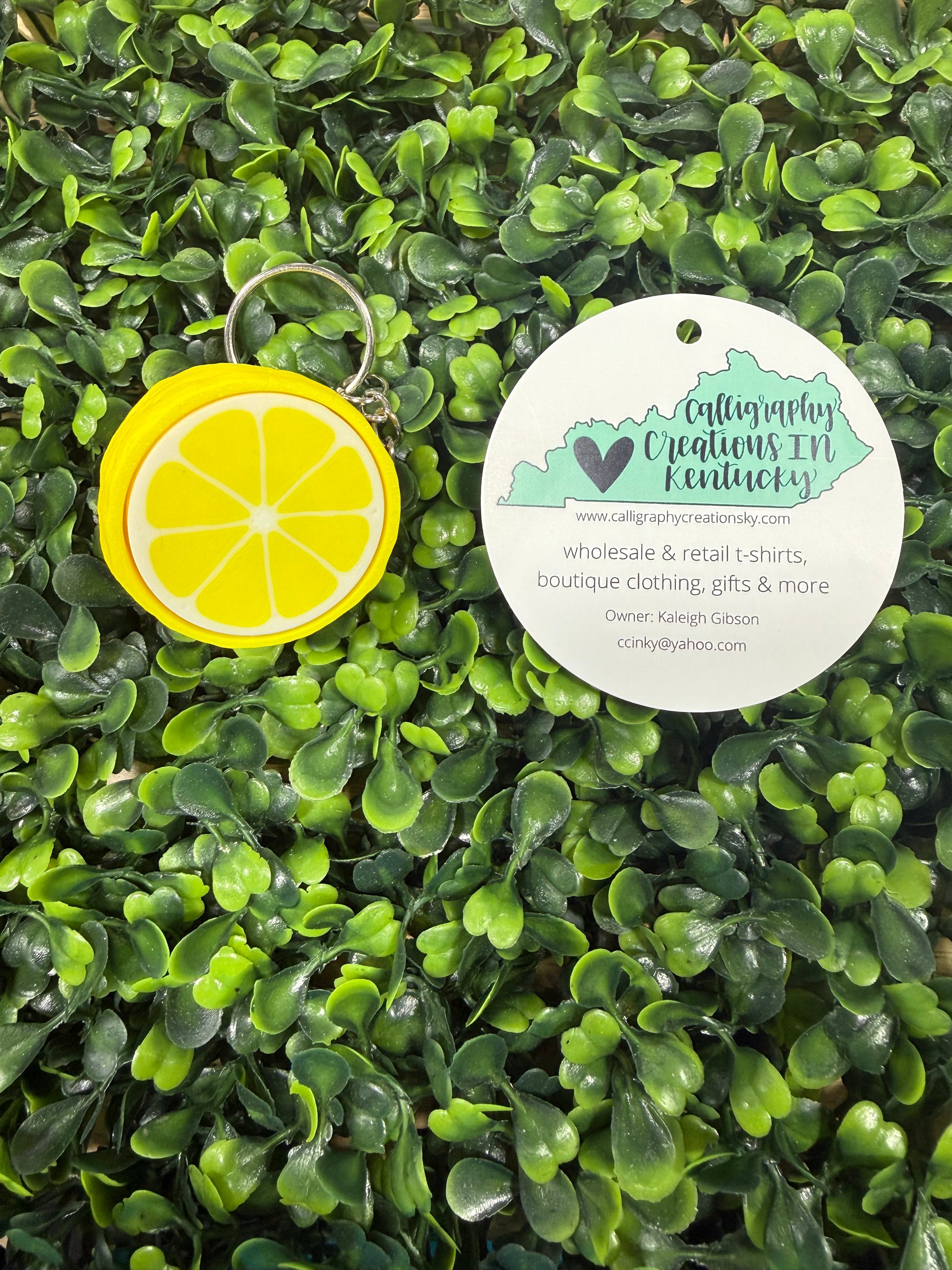 Yellow lemon-shaped keychain on a green leafy background with a circular tag.