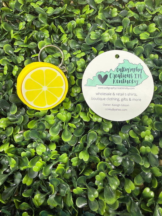Yellow lemon-shaped keychain on a green leafy background with a circular tag.