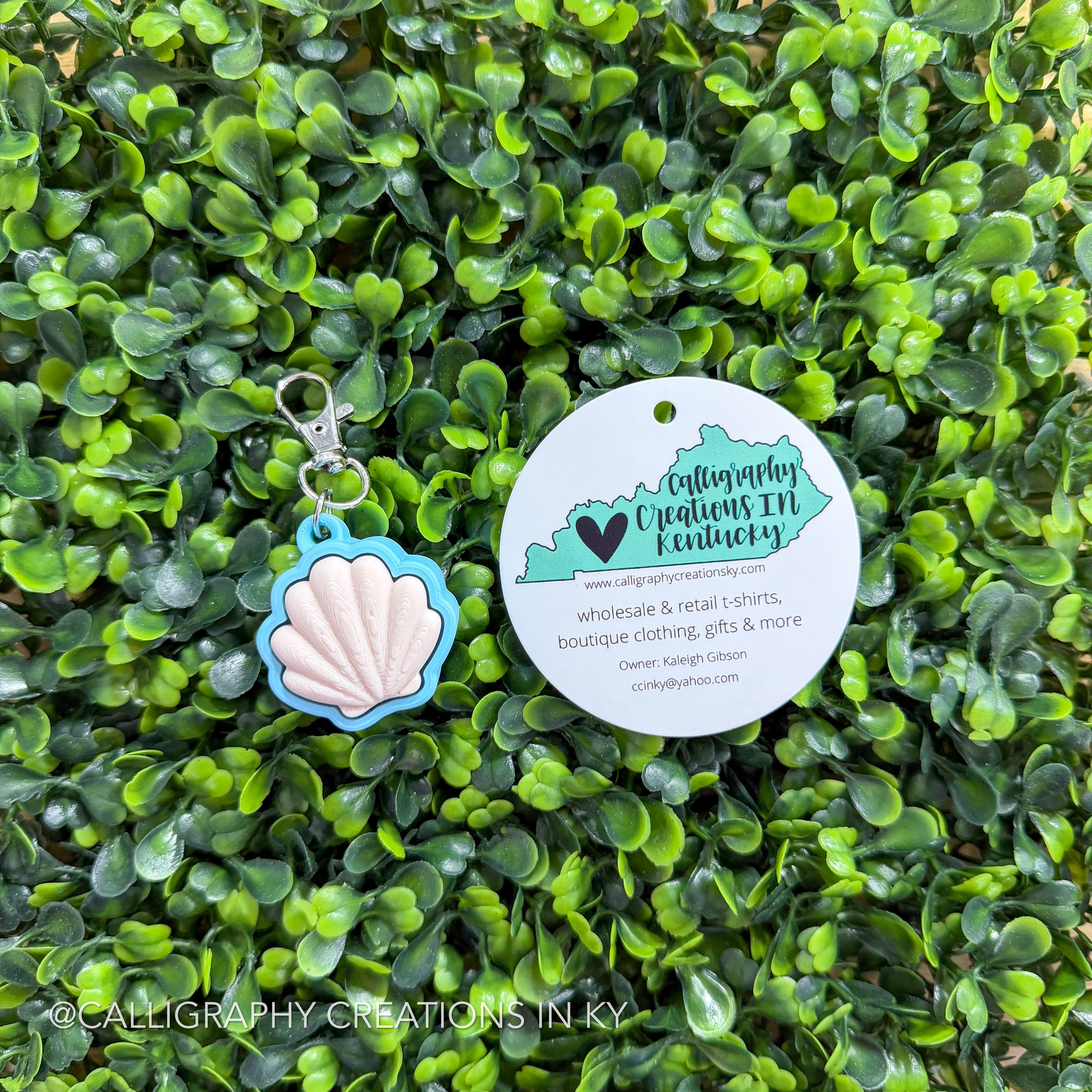 Shell-shaped fidget clicker keychain and round tag on a green leafy background with 'Calligraphy Creations in KY' branding.