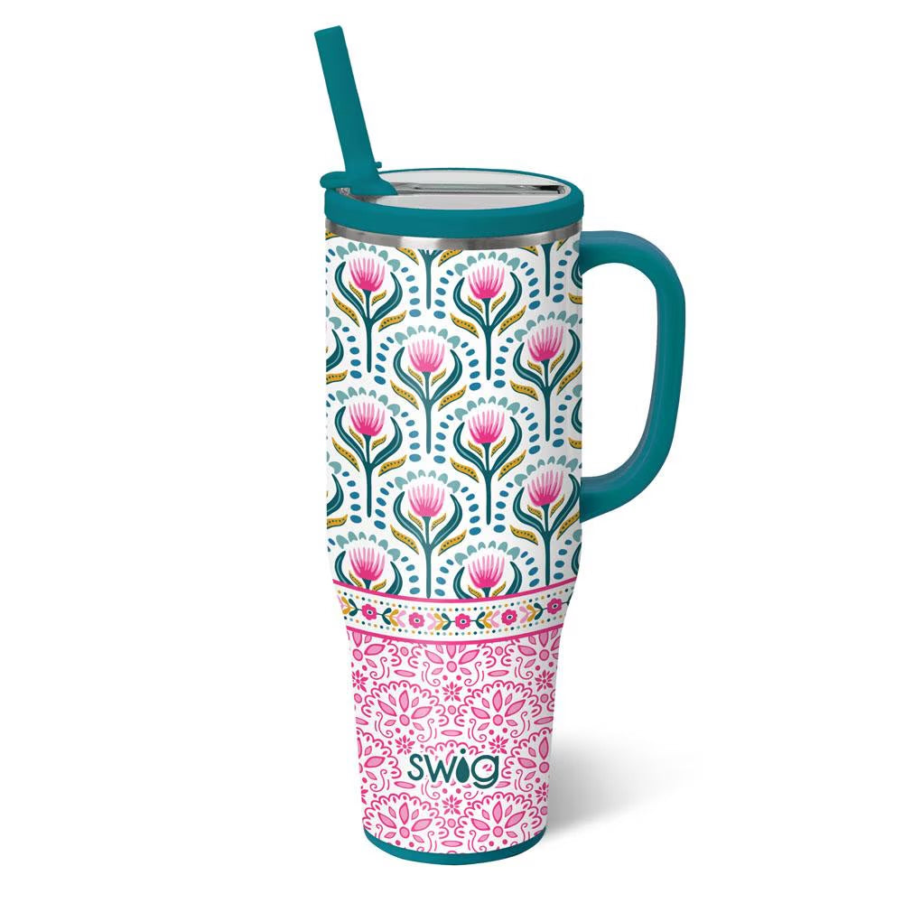 A 40 oz handled travel tumbler with a teal handle and straw, featuring a split design: the upper half has a white background with a colorful floral pattern, and the lower half is pink with a floral damask pattern. The brand name 'swig' is printed near the bottom.