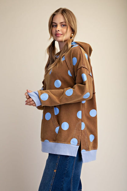 A woman modeling a brown hooded sweatshirt with large light blue polka dots and light blue ribbed cuffs and hem, paired with blue jeans against a plain beige studio background.
