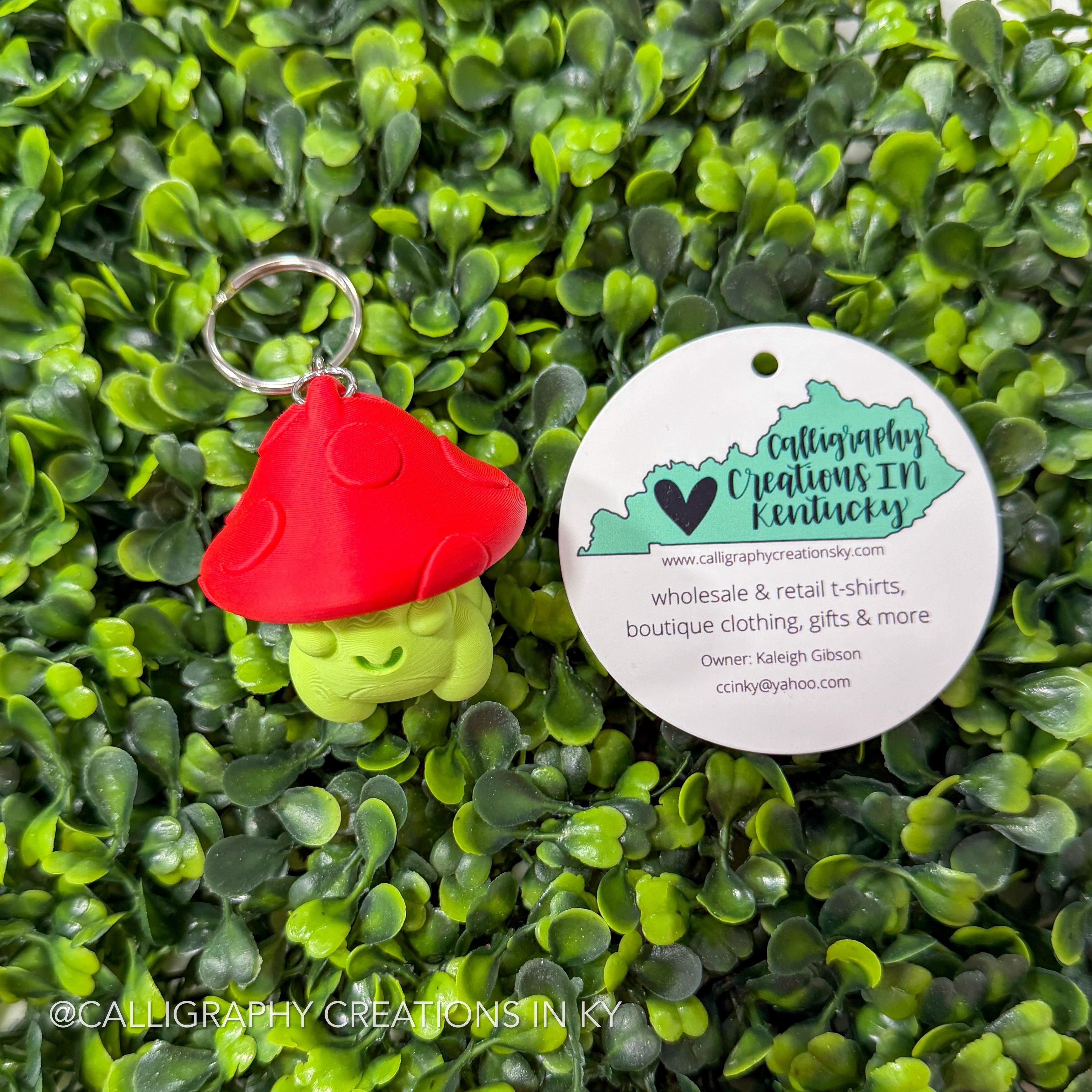 Red and green mushroom frog-shaped fidget clicker keychain with a 'Calligraphy Creations in Kentucky' tag on a green leafy background.