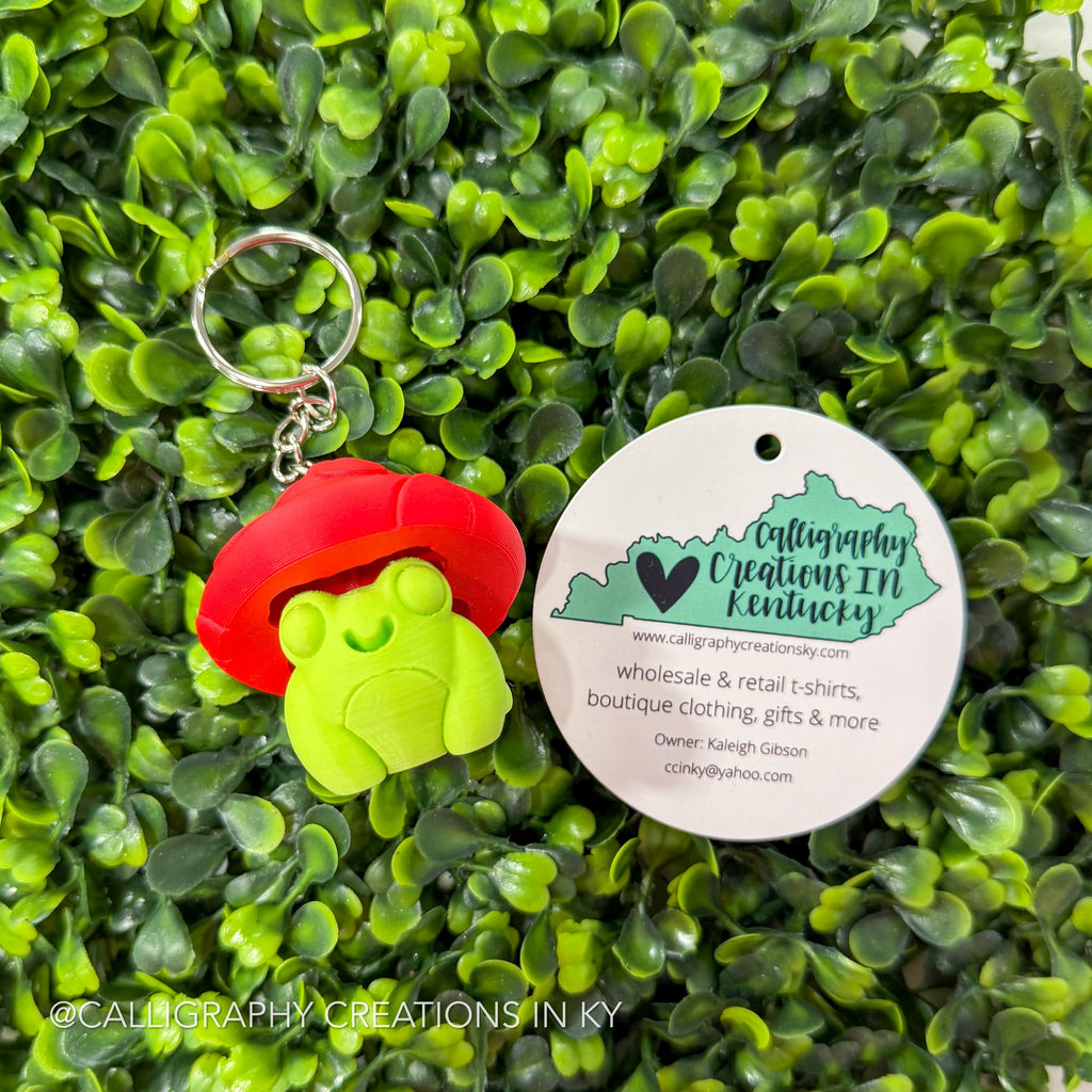 Fidget clicker Keychain with green frog and red mushroom hat on a green leafy background with a Calligraphy Creations In Ky brand tag.