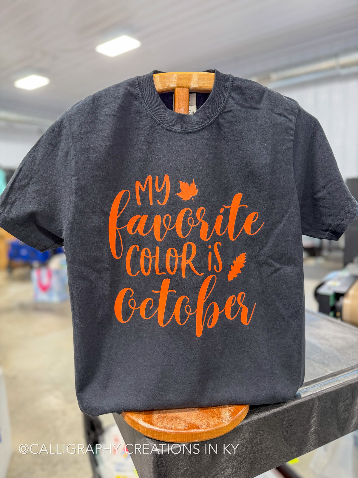 A dark charcoal gray t-shirt hanging on a wooden hanger, featuring the phrase 'MY favorite COLOR is October' in orange cursive script with small orange autumn leaf graphics. The background shows a blurred indoor retail or workshop environment.