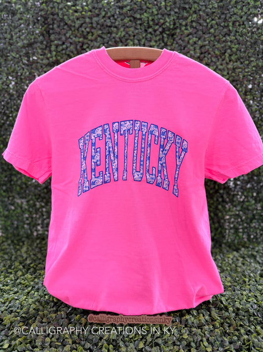 Neon pink shirt with blue floral letters spelling out ‘Kentucky’ on a wooden stand 