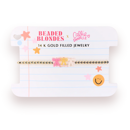 A 14K gold filled beaded bracelet featuring a pink bow and a small beige floral charm, presented on a white lined card with 'Beaded Blondes x Callie Danielle' branding.