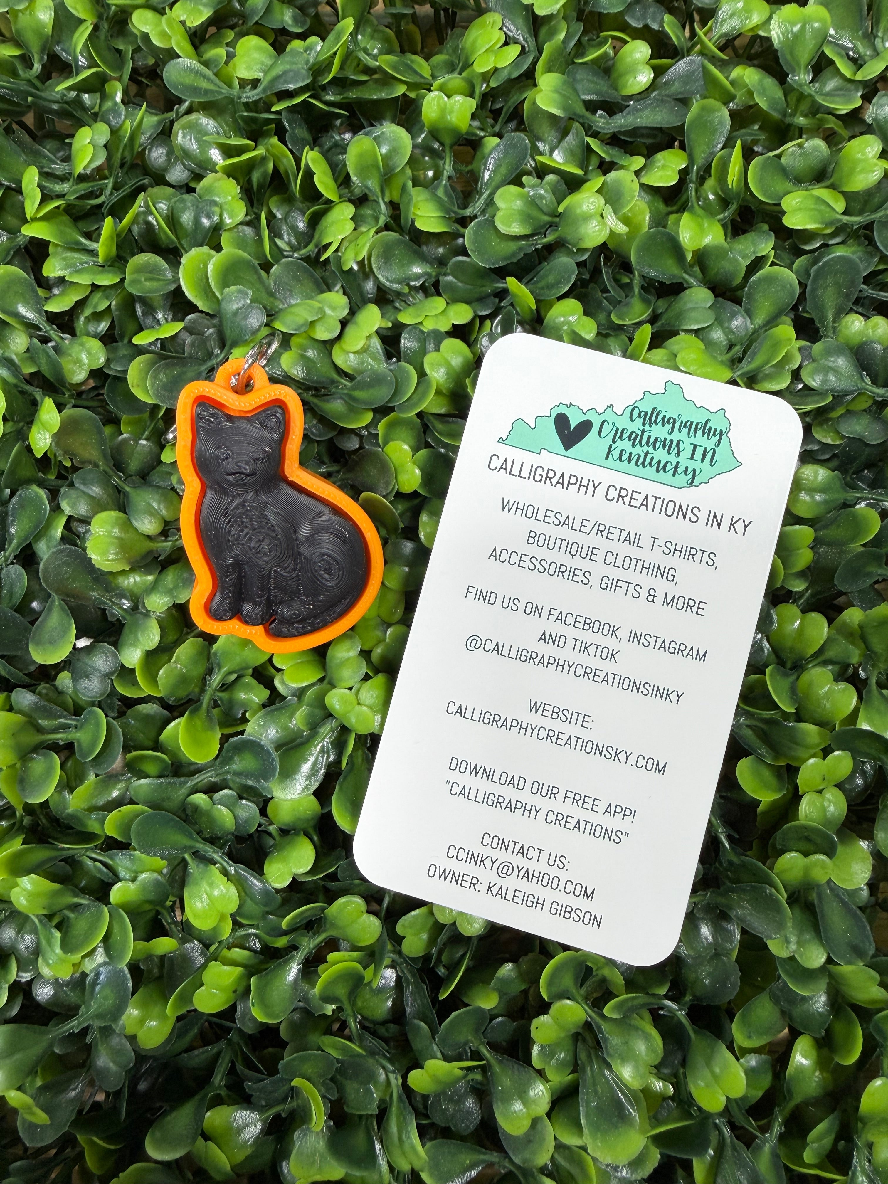 A black cat silhouette pendant with an orange border and a small metal loop, placed on green foliage next to a white product tag for Calligraphy Creations in KY.