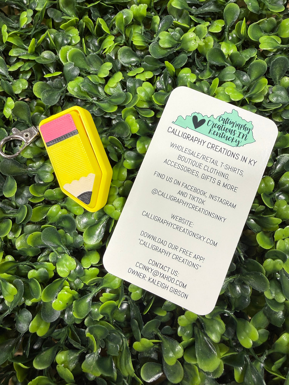 A yellow pencil-shaped keychain with a pink and yellow eraser lies on a white card printed with the brand name 'Calligraphic Creations in KY' over a dense green leaf background.