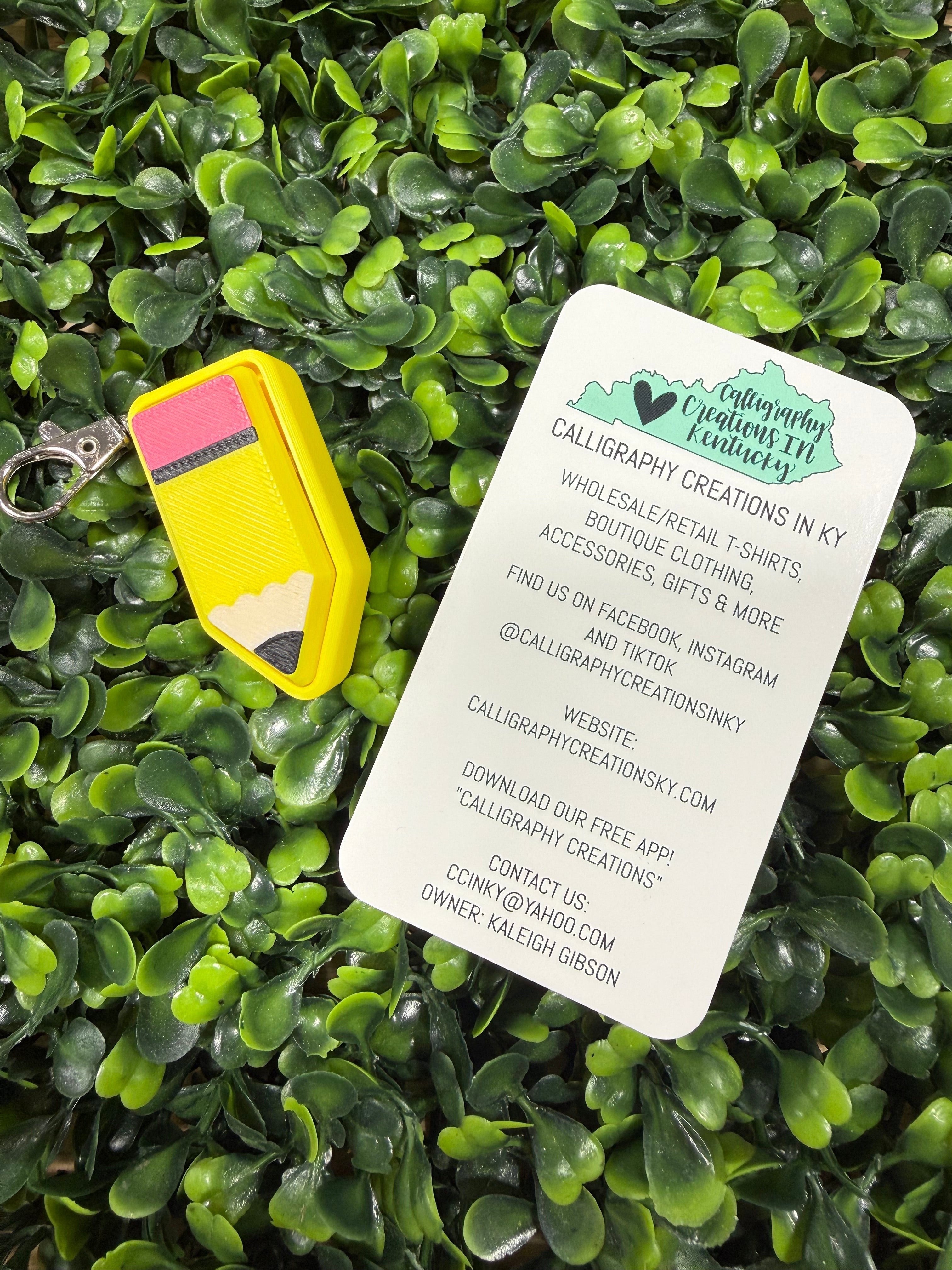A yellow pencil-shaped keychain with a pink and yellow eraser lies on a white card printed with the brand name 'Calligraphic Creations in KY' over a dense green leaf background.