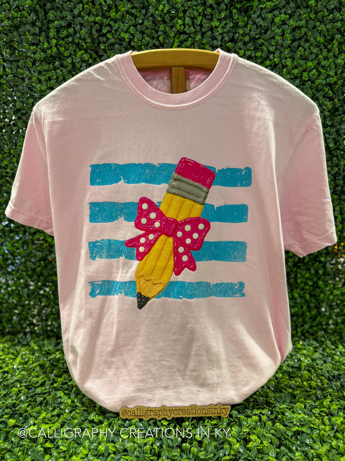 A light pink short-sleeve t-shirt on a wooden hanger, featuring a graphic of a yellow pencil with a pink polka-dot bow and striped background, set against a green hedge backdrop.