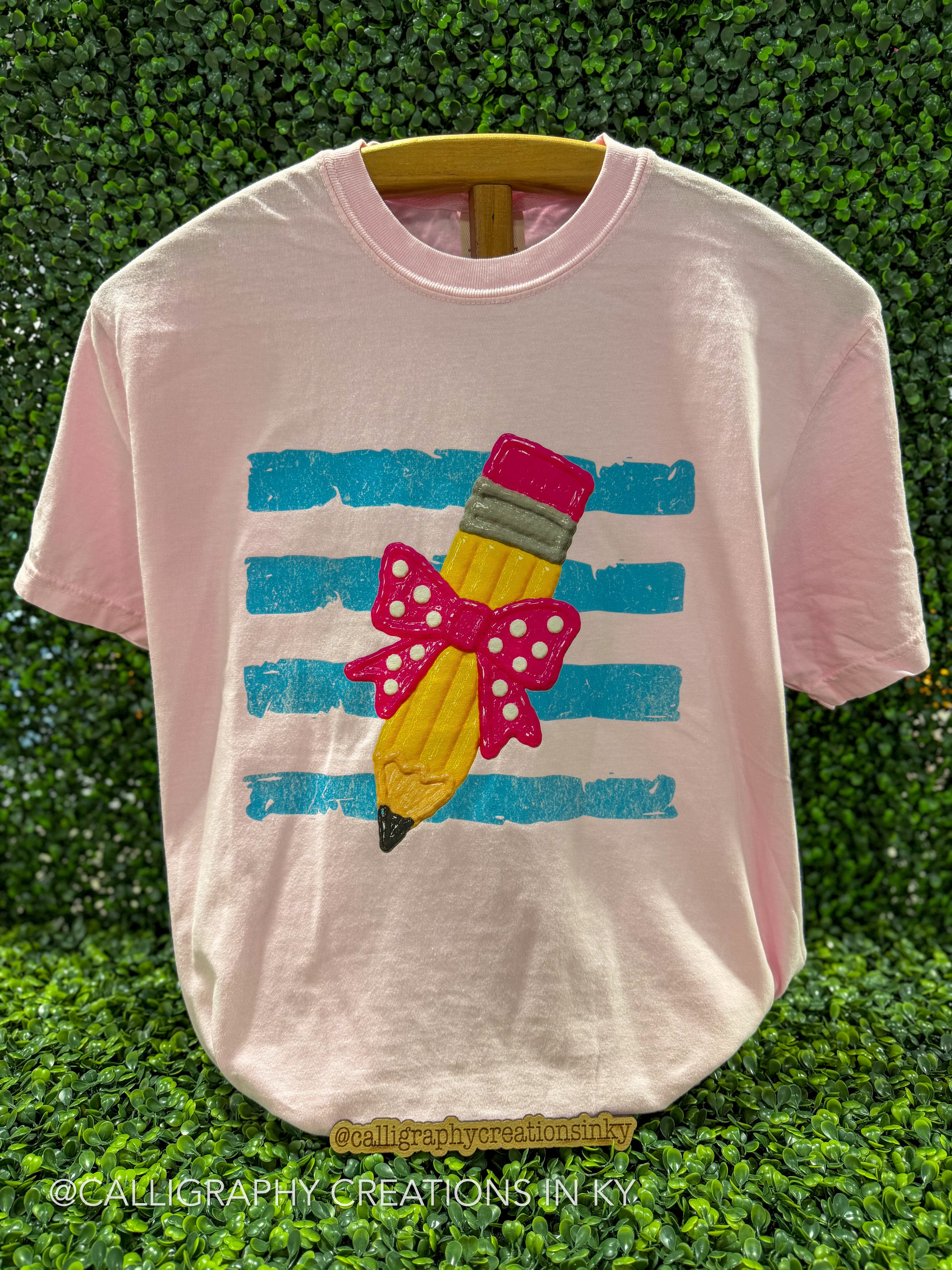A light pink short-sleeve t-shirt on a wooden hanger, featuring a graphic of a yellow pencil with a pink polka-dot bow and striped background, set against a green hedge backdrop.