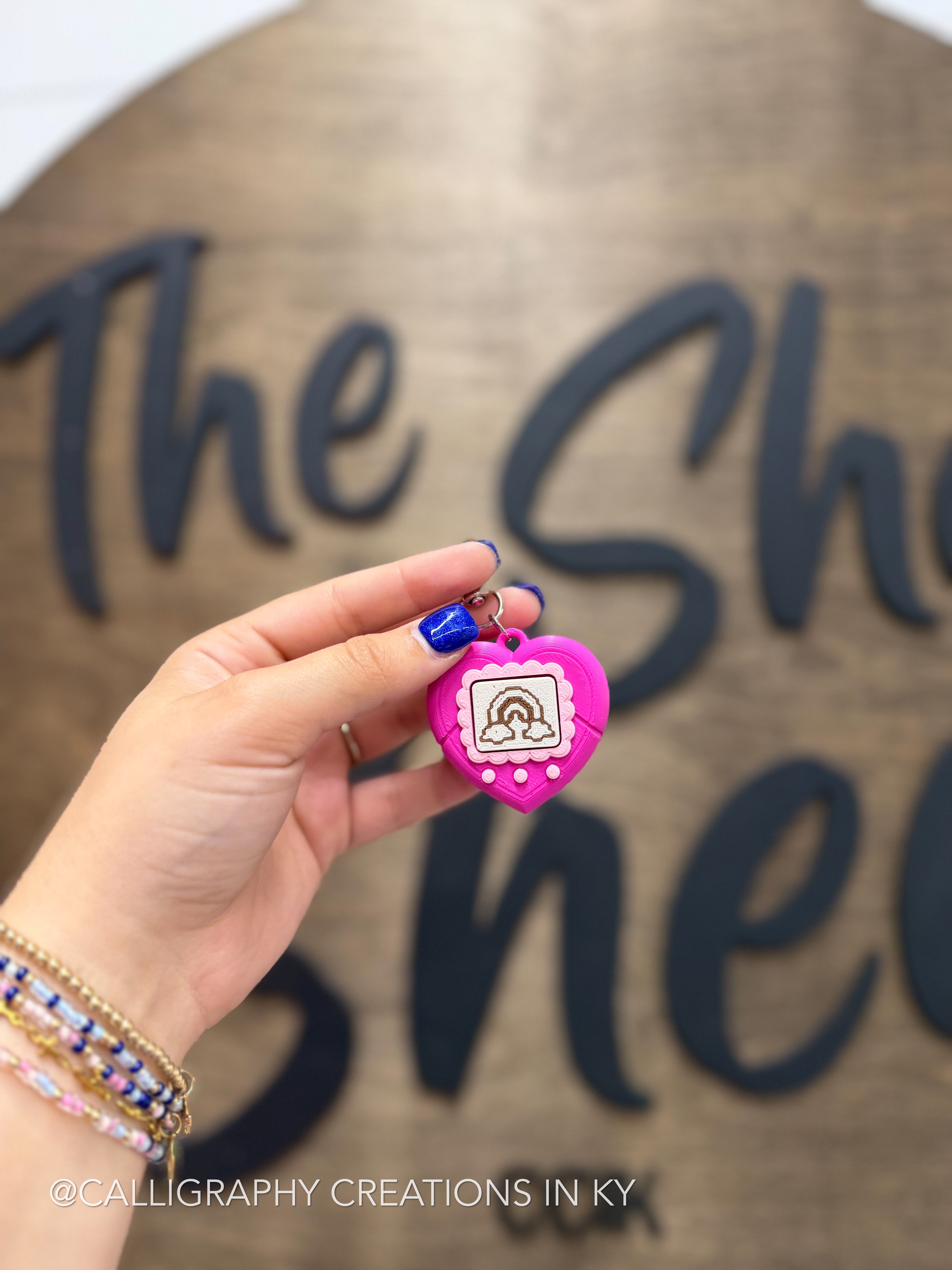 A hand holds a small pink heart-shaped 3d printed charm with a pink border, a rainbow design, and a metal loop, in front of a wooden sign with dark lettering.