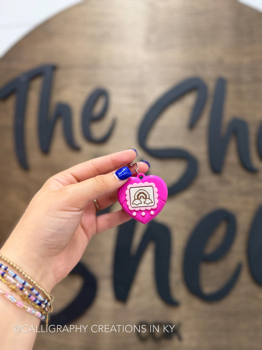 A hand holds a small pink heart-shaped 3d printed charm with a pink border, a rainbow design, and a metal loop, in front of a wooden sign with dark lettering.