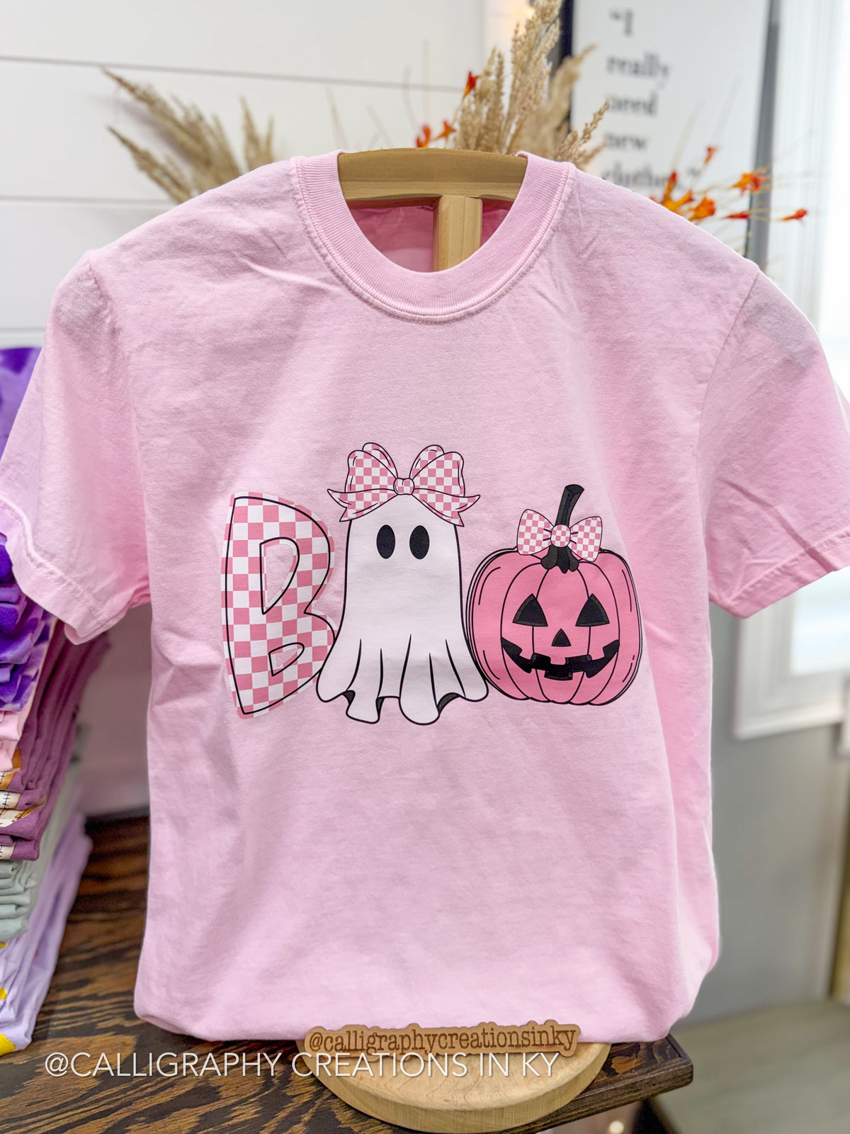 A light pink short-sleeve t-shirt displayed on a wooden hanger in a retail setting. The front features a Halloween-themed graphic spelling 'Boo' with a pink-and-white checkered 'B', a white ghost with a pink-and-white checkered bow, and a pink jack-o'-lantern pumpkin with a matching bow.