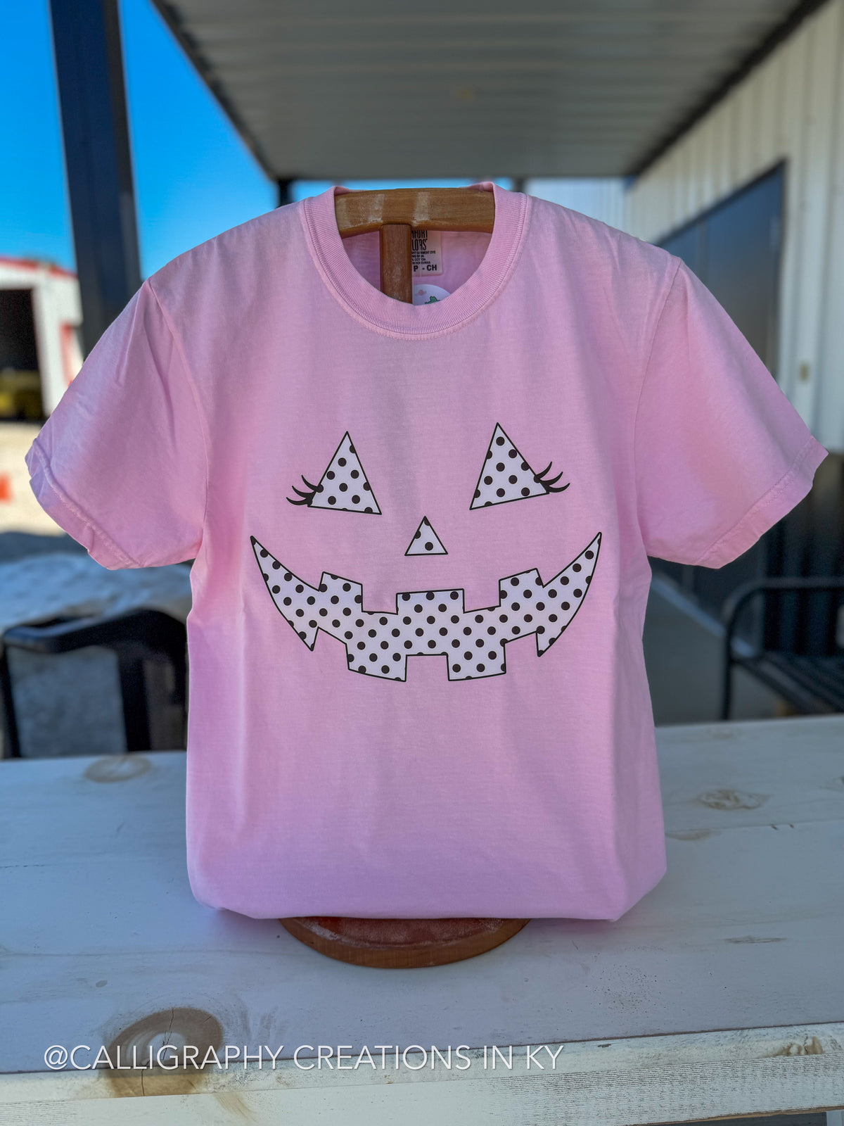 A light pink short-sleeve t-shirt displayed on a wooden hanger stand on a white wooden table. The shirt features a jack-o'-lantern face graphic made from black and white polka-dot triangles.