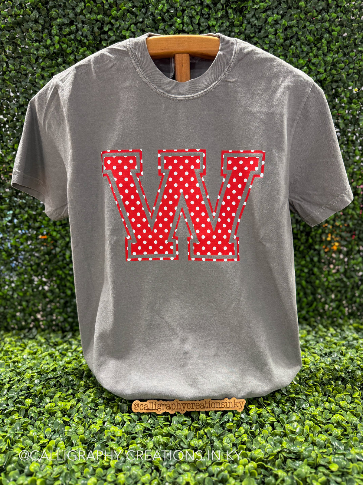 A gray short-sleeve t-shirt displayed on a wooden hanger against a green hedge background. The shirt features a large, stylized letter 'W' on the chest, filled with a red and white polka dot pattern and outlined with a dashed red and green stitched design effect.