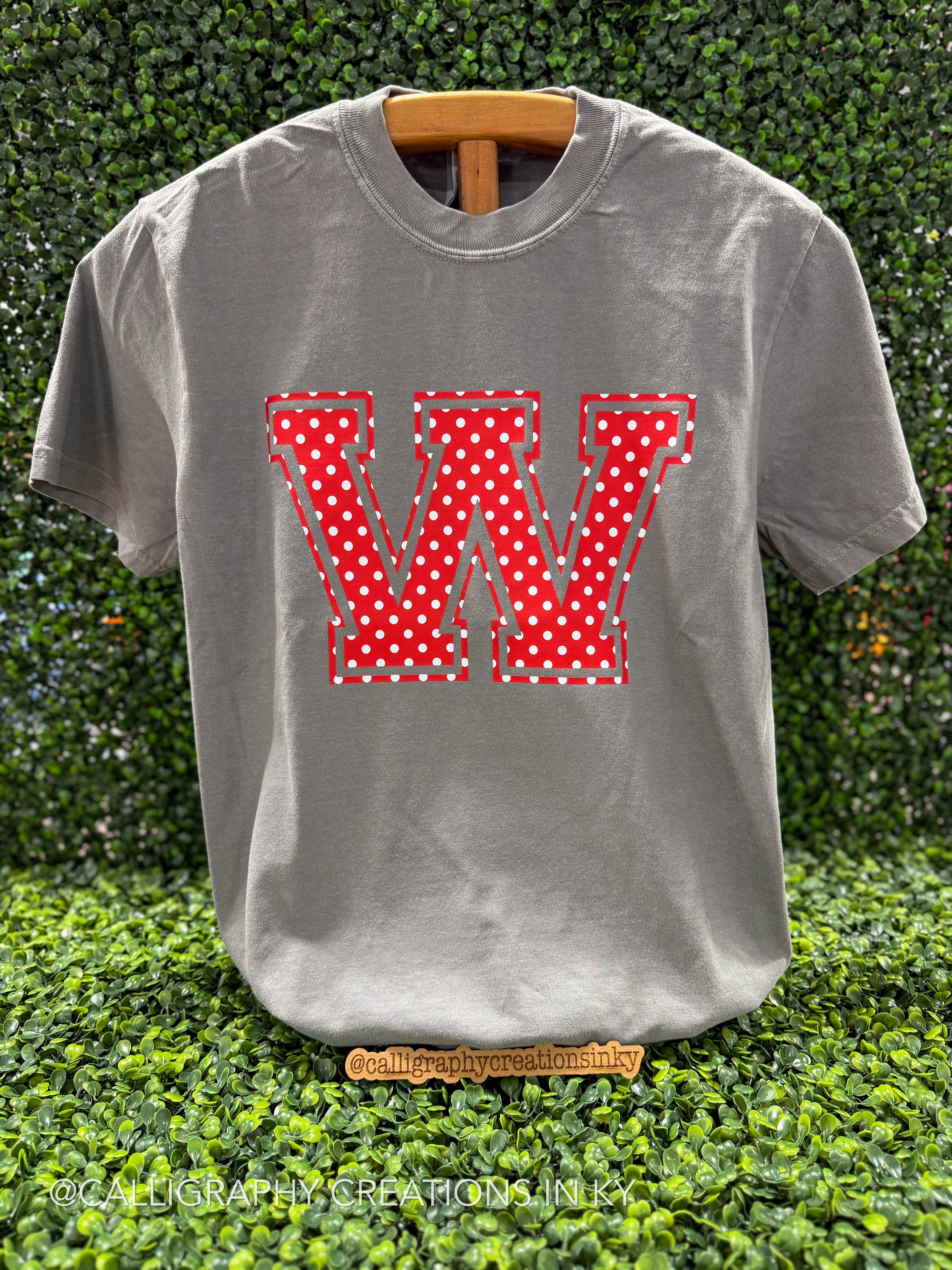 A gray short-sleeve t-shirt displayed on a wooden hanger against a green hedge background. The shirt features a large, stylized letter 'W' on the chest, filled with a red and white polka dot pattern and outlined with a dashed red and green stitched design effect.