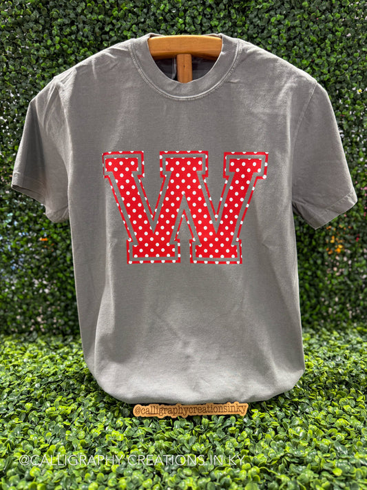 A gray short-sleeve t-shirt displayed on a wooden hanger against a green hedge background. The shirt features a large, stylized letter 'W' on the chest, filled with a red and white polka dot pattern and outlined with a dashed red and green stitched design effect.