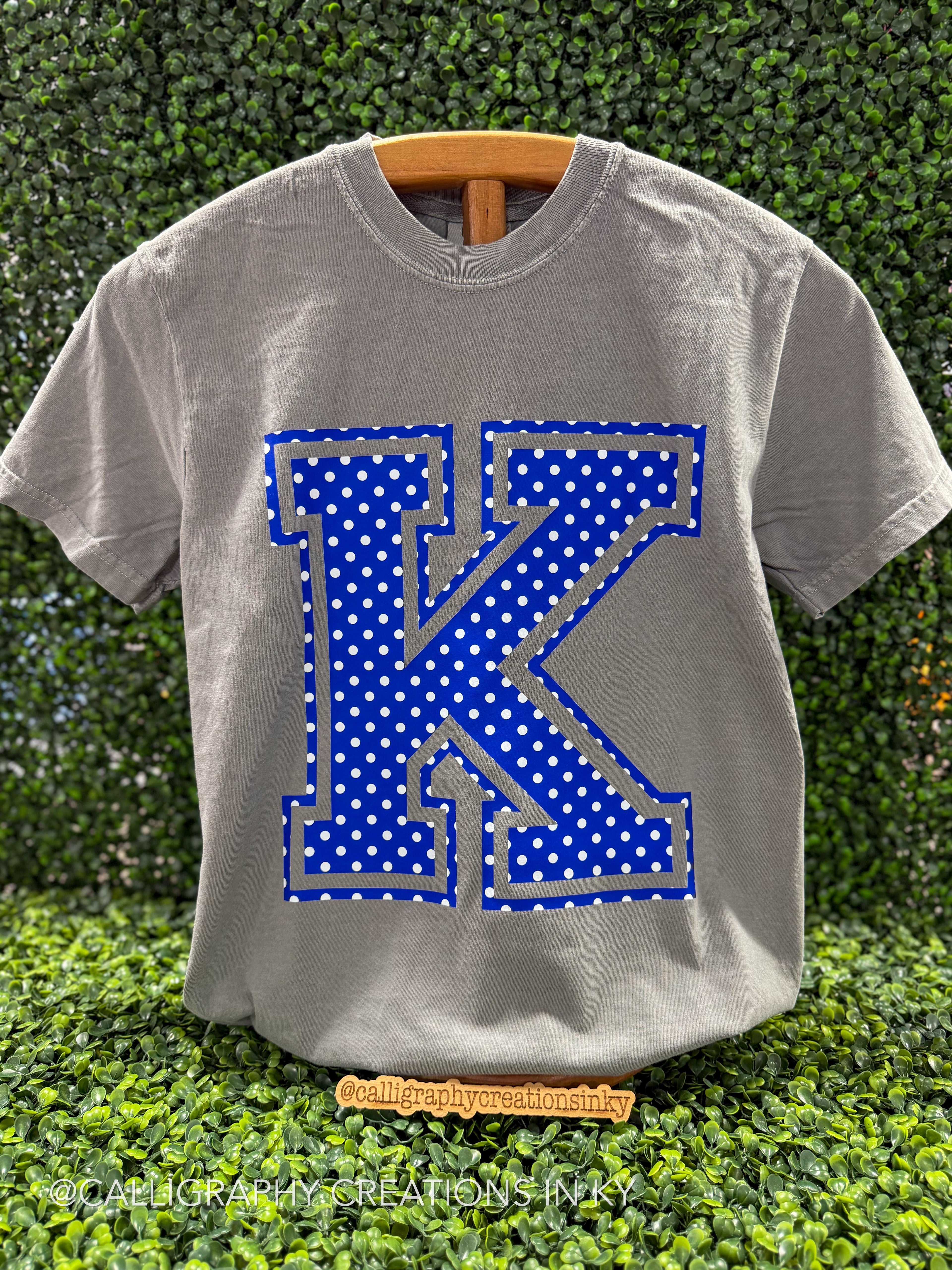A heather gray short-sleeve t-shirt on a wooden hanger, featuring a large blue and white polka dot letter 'K' graphic on the front, set against a green hedge background.