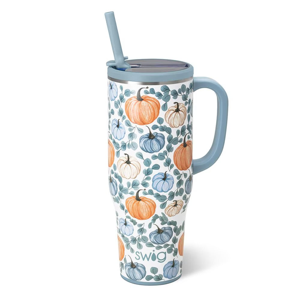 A tall insulated travel tumbler with a handle and straw, featuring a white background with an all-over pattern of orange, blue, and cream pumpkins and green leaves. The tumbler has a light blue lid and handle, and the brand name 'swig' is printed near the bottom.