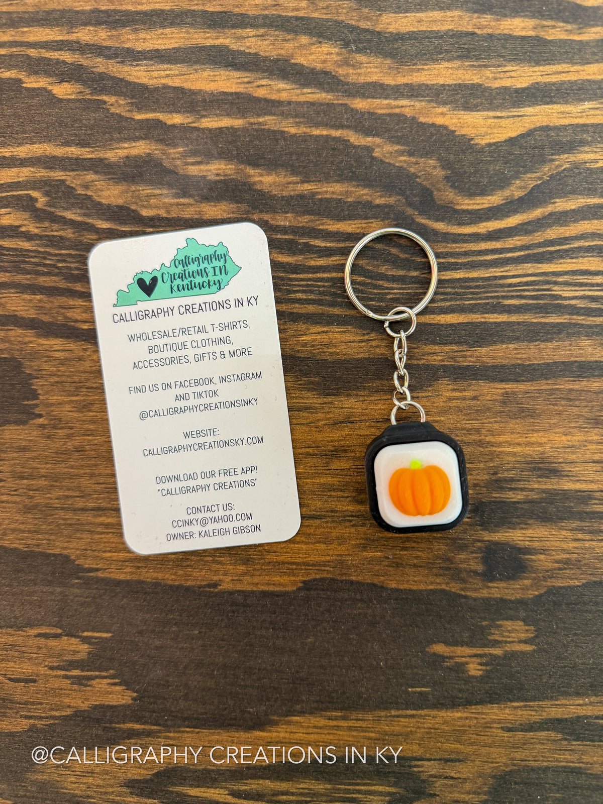 A small keychain with a square charm shaped like a pumpkin inside a black square, shown next to a white branded card on a dark wood surface.