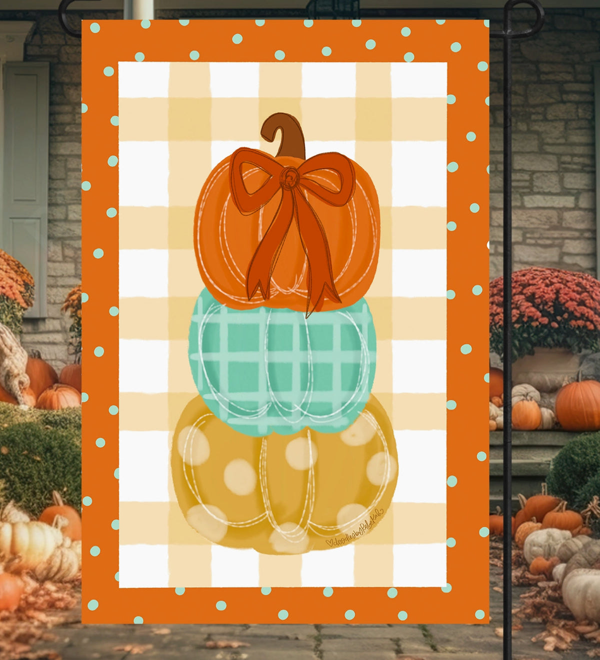 A vertical decorative garden flag displayed outdoors on a black metal stand. The flag features three stacked pumpkins in orange, mint green, and mustard yellow, set against a white and orange gingham background with a turquoise polka-dot border.