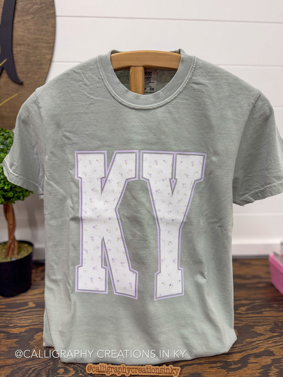 A light sage green short-sleeve t-shirt hanging on a wooden hanger, featuring a large white block letter 'K' and 'Y' outlined in lavender-purple with a tiny floral pattern inside.