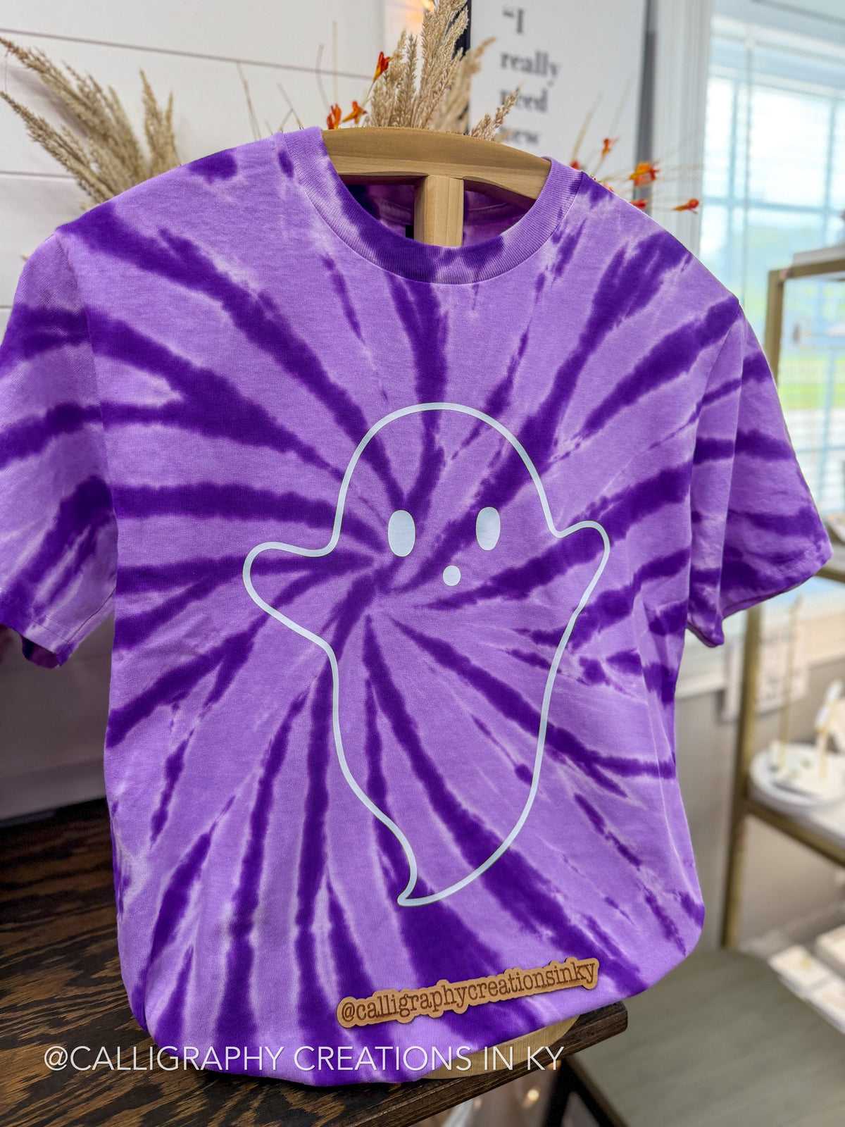 A purple and violet tie-dye t-shirt with a white ghost outline graphic on the front, hanging on a wooden hanger in a styled indoor setting.