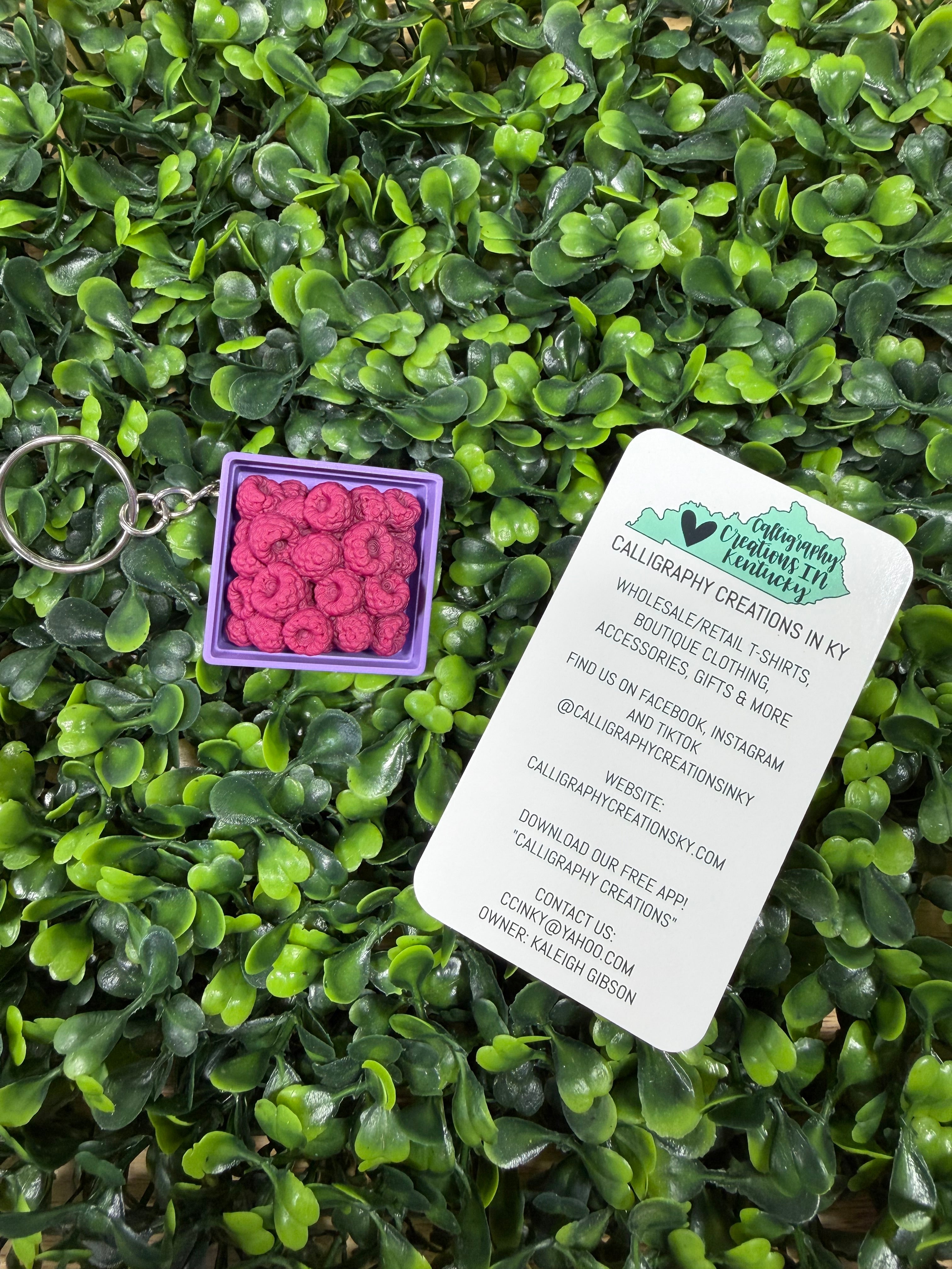 A small square purple box containing 3D printed raspberries keychain charms, placed on dense green foliage next to a white printed business card for Calligraphic Creations in KY.