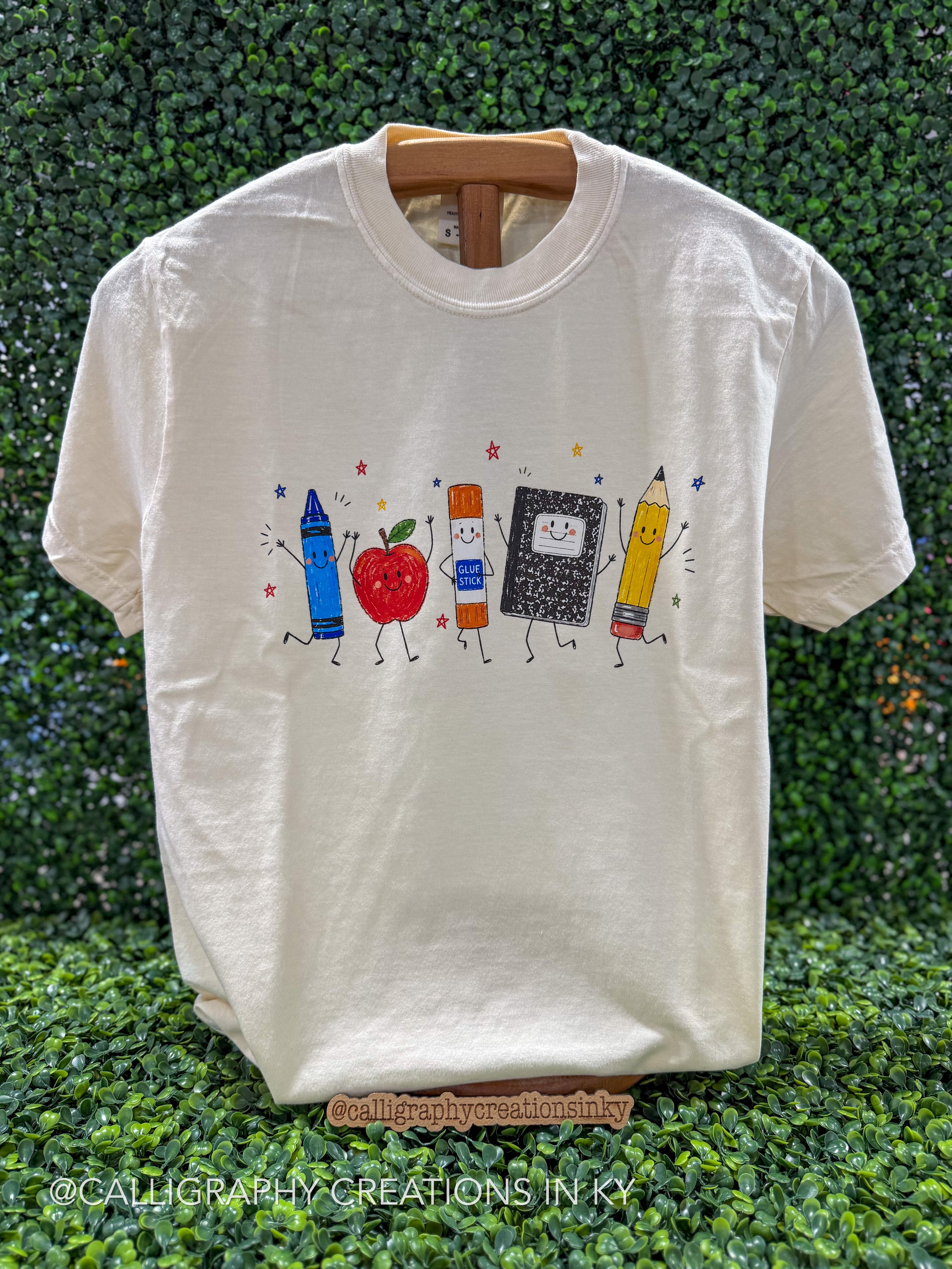 An off-white short-sleeve t-shirt on a wooden hanger, featuring a colorful cartoon graphic of school supplies (crayon, apple, glue stick, notebook, pencil) on the chest, set against a green hedge background.