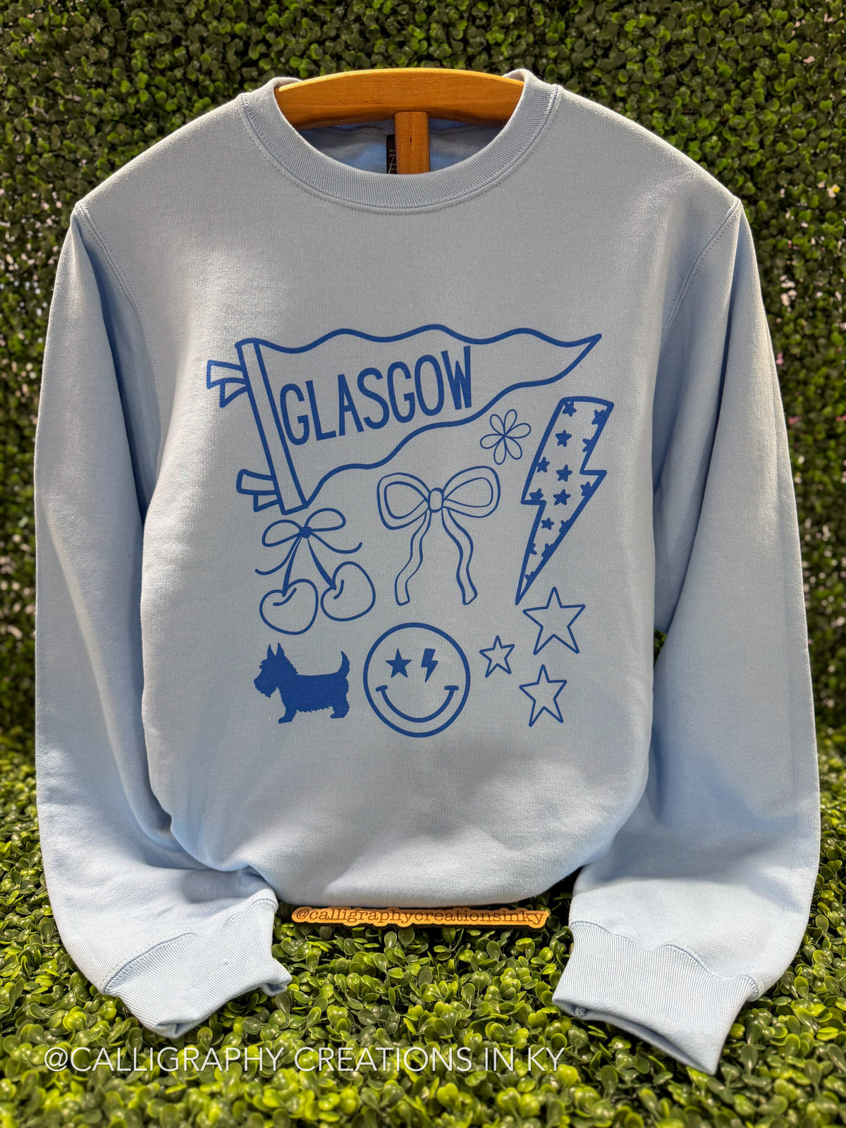 A light blue crewneck sweatshirt featuring a blue graphic print of a Glasgow flag, cherries, bow, lightning bolt, dog, smiley face, and stars, displayed on a wooden hanger against a green hedge background.
